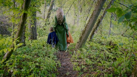 Two little toddler boys cosplay gnomes or hobbits with elf woman walking in green forest. Unrecognizable fairy tale characters. Halloween, kids concept. Amazing light. Slow motion. 
