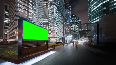 Modern billboard with a green screen on a busy street of the big city in the evening. Tall skyscrapers glittering with neon lights, road traffic and pedestrians passing by in motion timelapse.