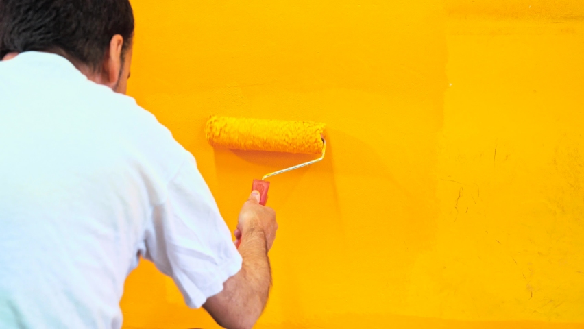 Painter man at work painting a orange wall with a roller, rear view. Offering professional painting services. 4k Stock video of professional painting services, contractor. Royalty-Free Stock Footage #1062895948