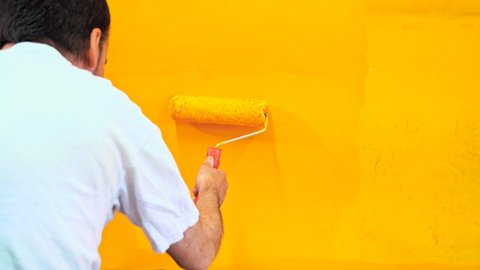 Painter man at work painting a orange wall with a roller, rear view. Offering professional painting services. 4k Stock video of professional painting services, contractor.