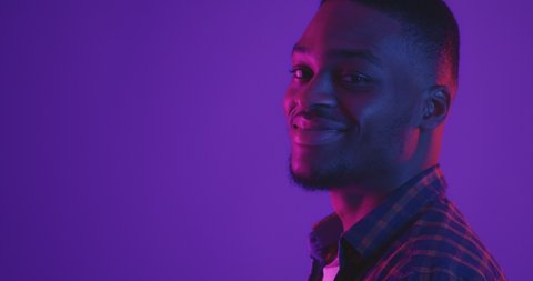 Side view portrait of young african american guy turning face to camera and laughing happily, neon lights, studio shot, empty space