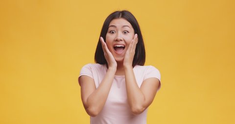 Unbelievable offer. Young surprised woman touching cheeks, opening mouth in amazement, expressing disbelieve and excitement, orange studio background