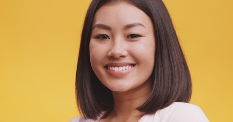Playful happy asian woman blinking eye, looking at camera with toothy smile, winking and flirting, expressing optimism, yellow studio background