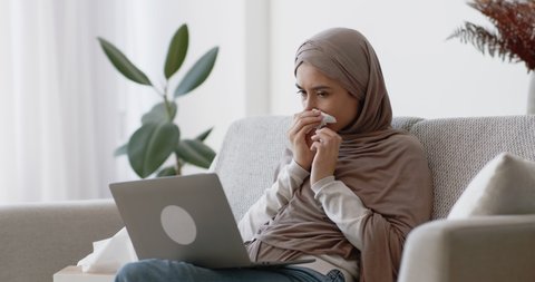 Sick muslim woman working on laptop at home, coughing and blowing her nose. Remote work and health care concept, slow motion