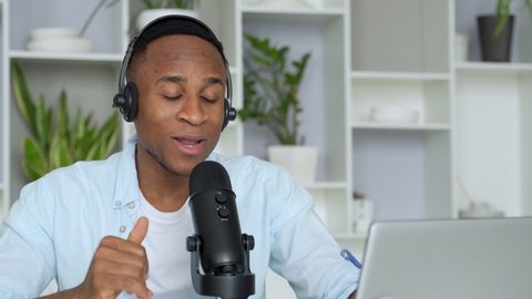 Podcast concept - happy young black man audio blogger in headphones with laptop computer and microphone broadcasting at home office