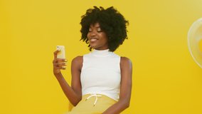 Young African woman is sitting in yellow room, relaxing, having online conversation with a colleague, using mobile phone, enjoying video call, creative video concept.
