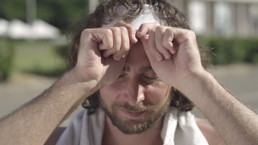 Close-up face of happy handsome Caucasian bearded man with brown eyes adjusting headband and smiling at camera. Portrait of positive confident sportsman posing in sunlight outdoors.