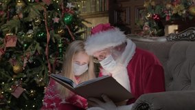 Santa Claus in medical mask visiting girl during coronavirus epidemic, Santa is reading magic book with bright light shining on them. Girl in period of coronavirus reading book with Santa.