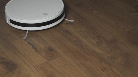 White robotic vacuum cleaner on laminate floor cleaning dust in living room interior.