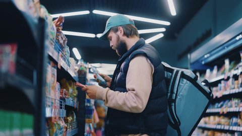 Careful young bearded courier man selecting food products for customer on shelves reading nutritional value information. Wise shopping. Online delivery. Jobs.