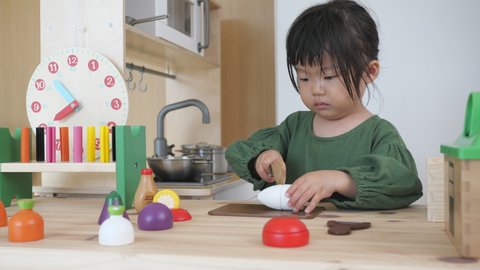 Asian 2 years old cute little toddler baby girl who cuts wooden toy radish with a knife in the play house kitchen. Put the vegetables in a toy steel pot