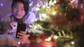 Asian female woman happiness cheerful hand holds smartphone video call to her friend lay down on the sofa with cute lovely puppy little dog in Christmas holiday vacation festive background concept.