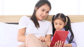 Asian mother and little girl using tablet for video call with their father working abroad in bedroom.