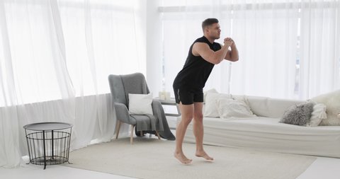 Home fitness, training, workout, bodycare and wellness concept. Sporty young man is doing lunge squat exercise in jump in living room at home. Athletic male is exercising, side view, camera in motion.