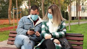 Young couple sitting on bench in park, wearing protective masks for COVID-19, watching video in cell-phone. Attractive blond woman and handsome man wearing medical mask outdoor, spending quality time