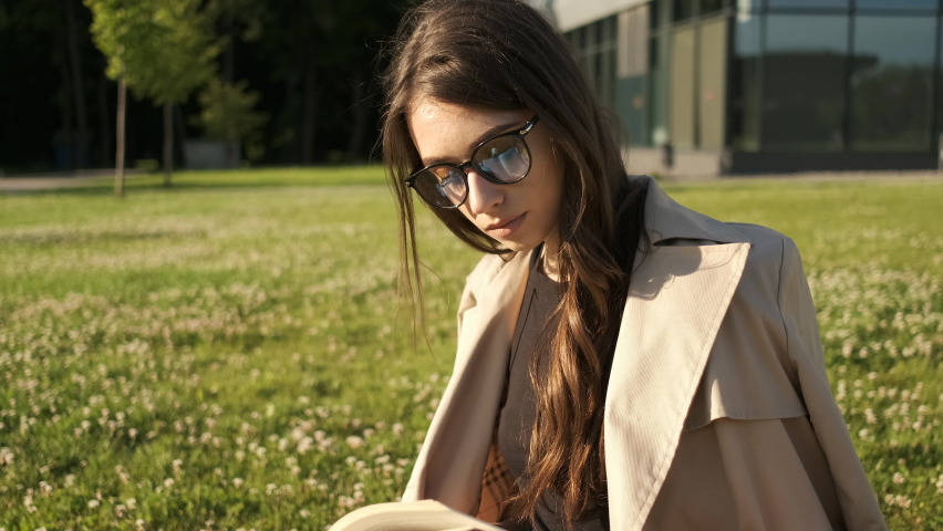 Stylish girl in glasses and a brown cloak sits in the park on the grass and reads a book. Student learning. Gift packages. Sunny summer evening Royalty-Free Stock Footage #1062938038