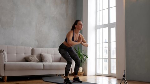 Fit athletic girl is engaged in squats and online lessons, exercises on a smartphone. Woman doing sports and fitness at home during covid quarantine. Healthy lifestyle during a pandemic