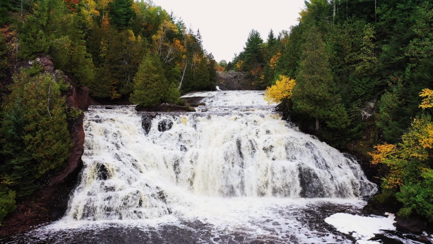 A closeup aerial of Lower Potato River Falls with heavy water flow cascading down the rocky stepped cliffs to the river below with trees lining the shore.