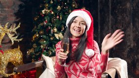 Portrait of a young happy woman in santa hat holding a glass of champagne and sending an air kiss while recording a video congratulation at home