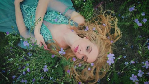  fairy tale sleeping beauty. Fantasy woman lies on blooming meadow in long blue medieval vintage dress eyes closed. Summer nature, green grass, bed from purple flowers. Blonde Girl enchanted princess