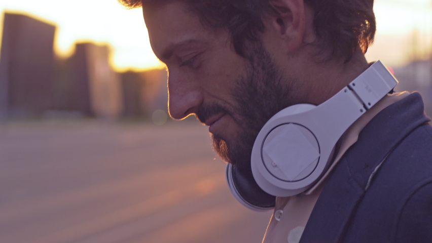 Young man enjoying music on the urban city street. Close-up of smiling male putting headphones on and listening to favorite song at sunset with lens flare. Royalty-Free Stock Footage #1062950554