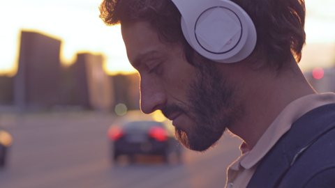 Young man enjoying music on the urban city street. Close-up of smiling male putting headphones on and listening to favorite song at sunset with lens flare.
