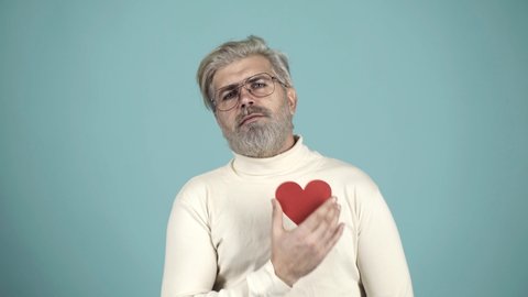 Heart pain. Broken heart concept. Portrait of grimacing sad upset nervous unhappy frustrated exhausted bearded man with heart shape isolated on gray blue background.