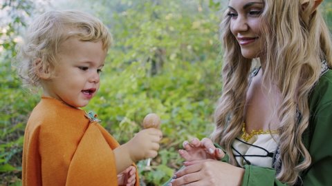 Fairy elf cosplay young woman treats little babies boys hobbits with candies in green forest. Halloween concept, fairytale characters, kids.