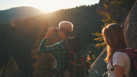 Mixed race young active couple hikers watching in binoculars at sunsets enjoying evening landscape on mountain travel in summertime. Activity concept.
