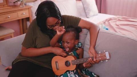 Close up African American mother play ukulele with her child on sofa in bedroom and they look enjoy and happy. Concept of new normal lifestyle and stay at home together.