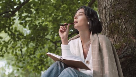 Lockdown of young pretty woman wearing casual clothes is sitting under the tree in park, writing in notebook then looking at camera and smiling