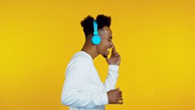 Handsome african man having fun, dancing with blue headphones in yellow studio. Side, profile view. Music, dance, radio concept, slow motion