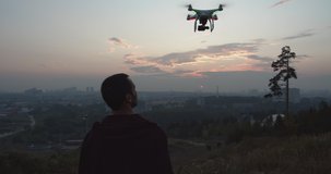 Silhouette of a man who stands on a hill against the background of the city stretches his hand up in order to catch a drone. High quality 4k footage.