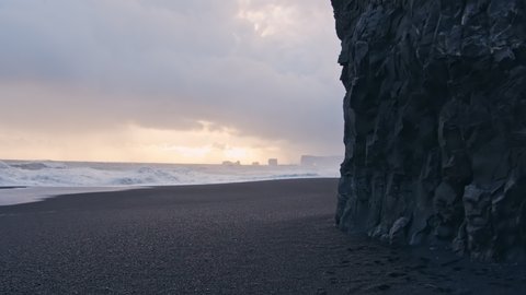 Wide Handheld Slow Motion Shot Of Black Sand Beach And Cliff Face With Crashing Surf Under Cloudy Sky At Sunset, Iceland