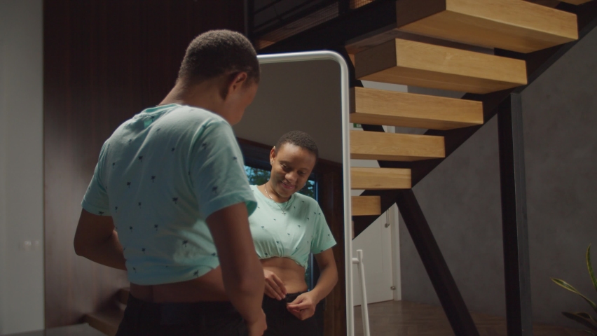 Happy joyful attractive african woman looking at her reflection in mirror, satisfied with her appearance after successful weight loss program in domestic room, expressing confidence and positivity.