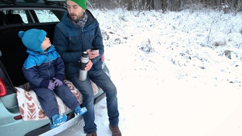 Winter road trip. Father and son sitting inside car trunk in snowy forest, drinking hot tea from thermos and sing a song. Outdoor picnic, family activity on nature in cold season.