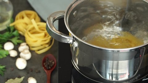 Male hands put Fettuccine pasta in a pan of boiling water and stir with a spoon on the background of mushrooms, greens and olive oil. Cooking pasta. Italian Cuisine. Close up.