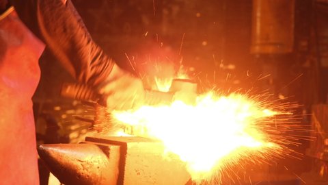 Medium shot of a man with a hammer hitting red-hot metal. A blacksmith works with metal in a forge. Sparks from impacts to metal