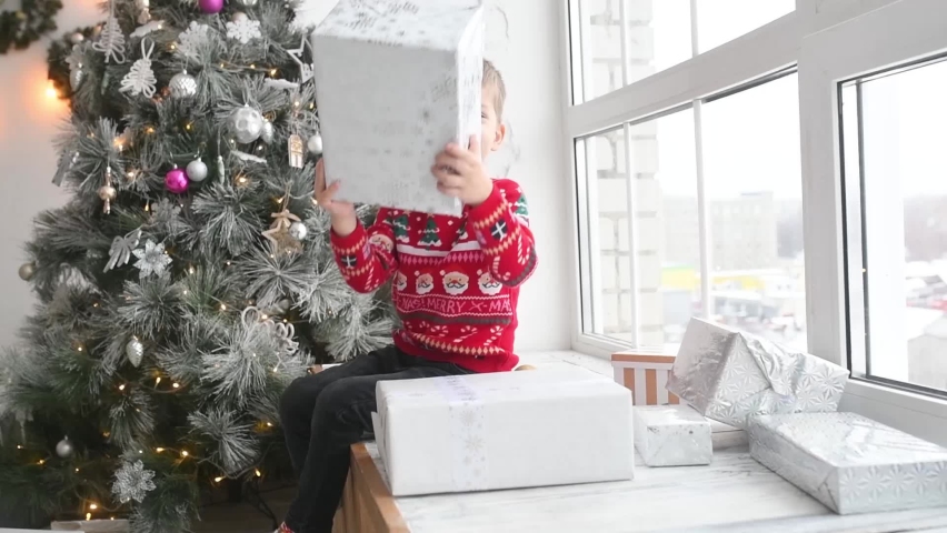 A little boy in a red sweater holds a gift in his hands, sitting on the windowsill, next to a Christmas tree and festive decorations