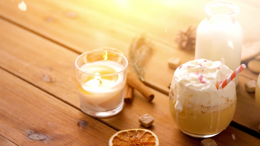 christmas, winter holidays and seasonal drinks concept - glasses of eggnog with whipped cream topping and paper straws, ingredients and candle burning on wooden background over snow falling