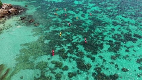 Aerial drone shot of red and yellow kayaks floating in crystal clear shallow water above coral reef in Thailand. Chasing camera flying parallel behind the group. Sunny summer weather, turquoise sea