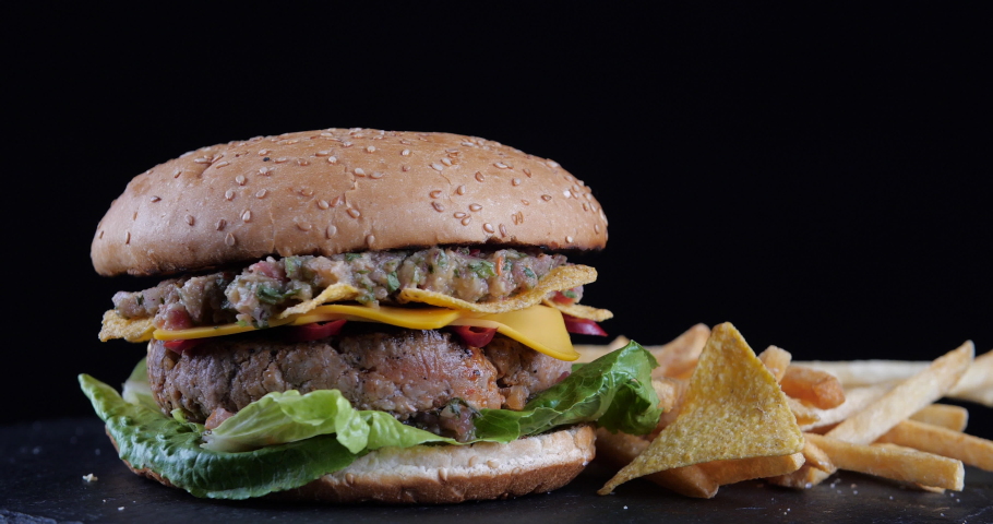 Close-up juicy fresh appetizing tasty burger hamburger chisburger lies on black table against dark background with fries and chips, fast food in diner pub restaurant, American dish, hunger concept