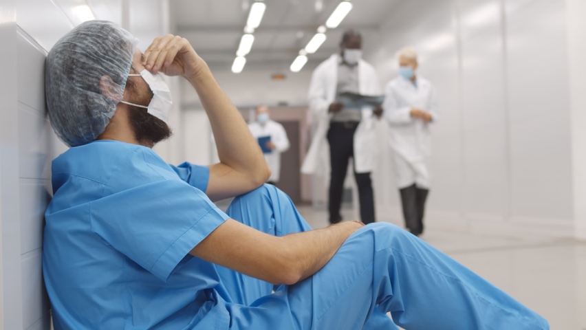 Colleagues supporting and cheering surgeon sitting on floor in hospital corridor. Doctors calming and helping tired and stressed medical worker in scrubs and safety mask resting on floor | Shutterstock HD Video #1062996319
