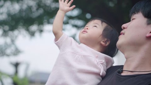 young Asian dad holding his baby girl looking up to the sky waving hand at park under trees, child care parent love bond, children innocence, father and child happy scene, hope for next generation