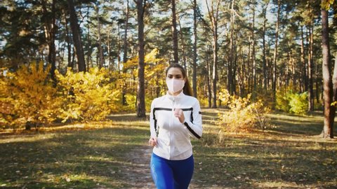 Young female in protective mask and sportswear is running by path in autumn wood. Sunny day, yellow trees. Healthy lifestyle. Covid-19, coronavirus worldwide pandemic. Tracking shot, slow motion