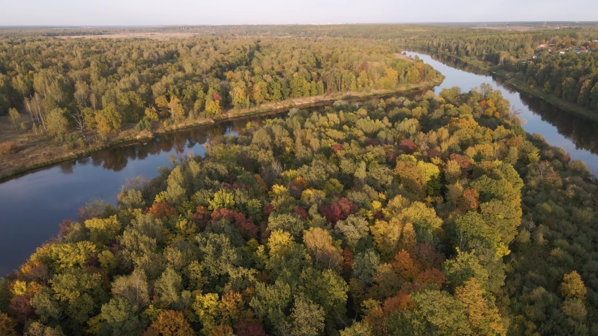 Flight over a dense forest standing on the study of the river. The leaves of the trees are colored yellow, orange and red. In the distance the sky at sunset. Autumn season. Soft evening light. 4K
