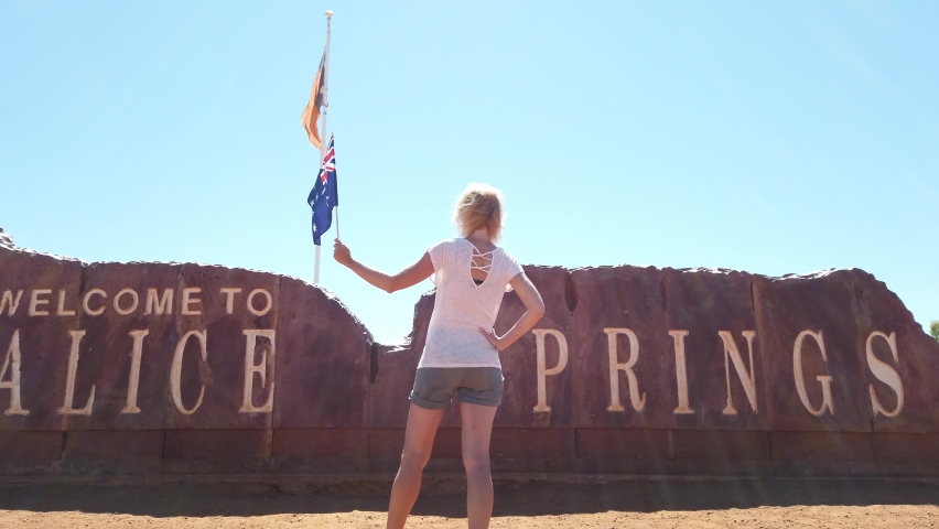 Happy carefree tourist woman at Alice Springs Welcome Sign in Northern Territory, Central Australia. Tourism in Outback Red Center desert. Travel discovery road in Australian trip in dry summer season