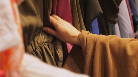 Manicured lady hands touch dress of brown colour among different coloured clothes in local shopping centre. Concept sales and discount