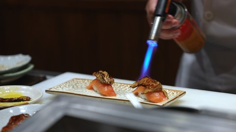 Close up of Professional chef hand use butane torch burner burning salmon sushi with foie gras on serving plate. Chef preparing healthy sushi menu on open kitchen counter bar in japanese restaurant.