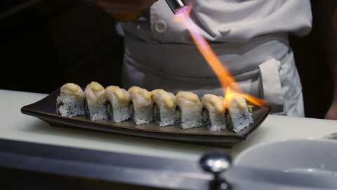 Close up of Professional chef hand use butane torch burner burning sushi roll with mayonnaise on serving plate. Chef preparing healthy sushi menu on open kitchen counter bar in japanese restaurant.